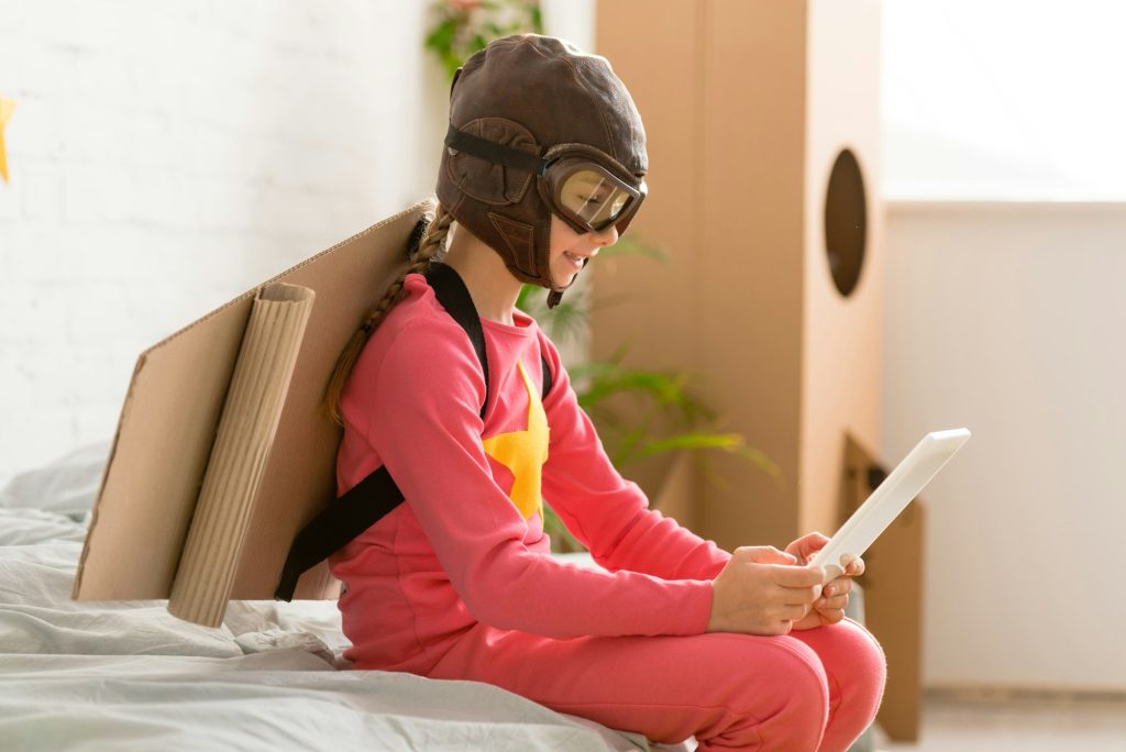 Adorable child with cardboard wings sitting on bed and using digital tablet